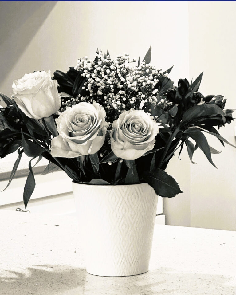 A black and white photo of a bouquet with three roses, babys breath, and greenery arranged in a patterned white vase, placed on a light-colored countertop.