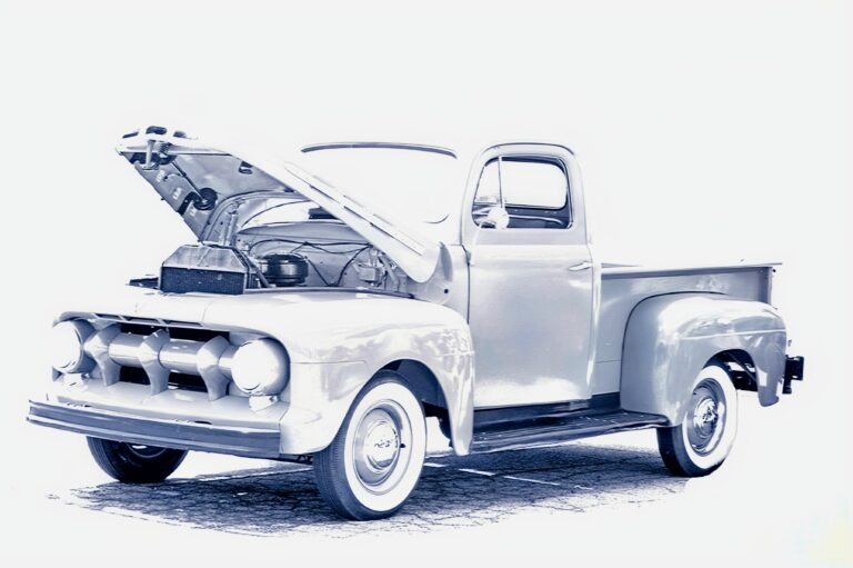 A vintage pickup truck with its hood open, revealing the engine, is parked on a cobblestone surface. The image has a high-contrast, faded effect, giving it a classic, nostalgic appearance.