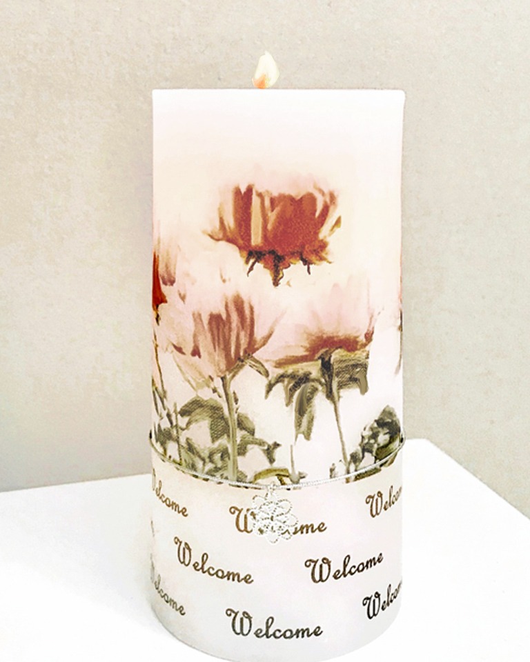 A lit white pillar candle decorated with pink flowers and a ribbon at the base that reads Welcome repeatedly, placed against a light-colored wall.