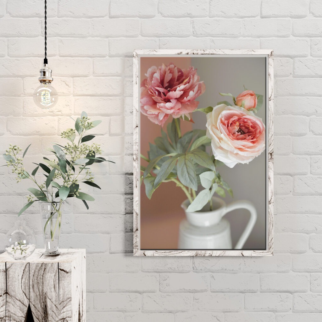 A framed photo of pink and white peonies in a jug hangs on a white brick wall, next to a hanging bulb and a vase with greenery on a rustic wooden table.